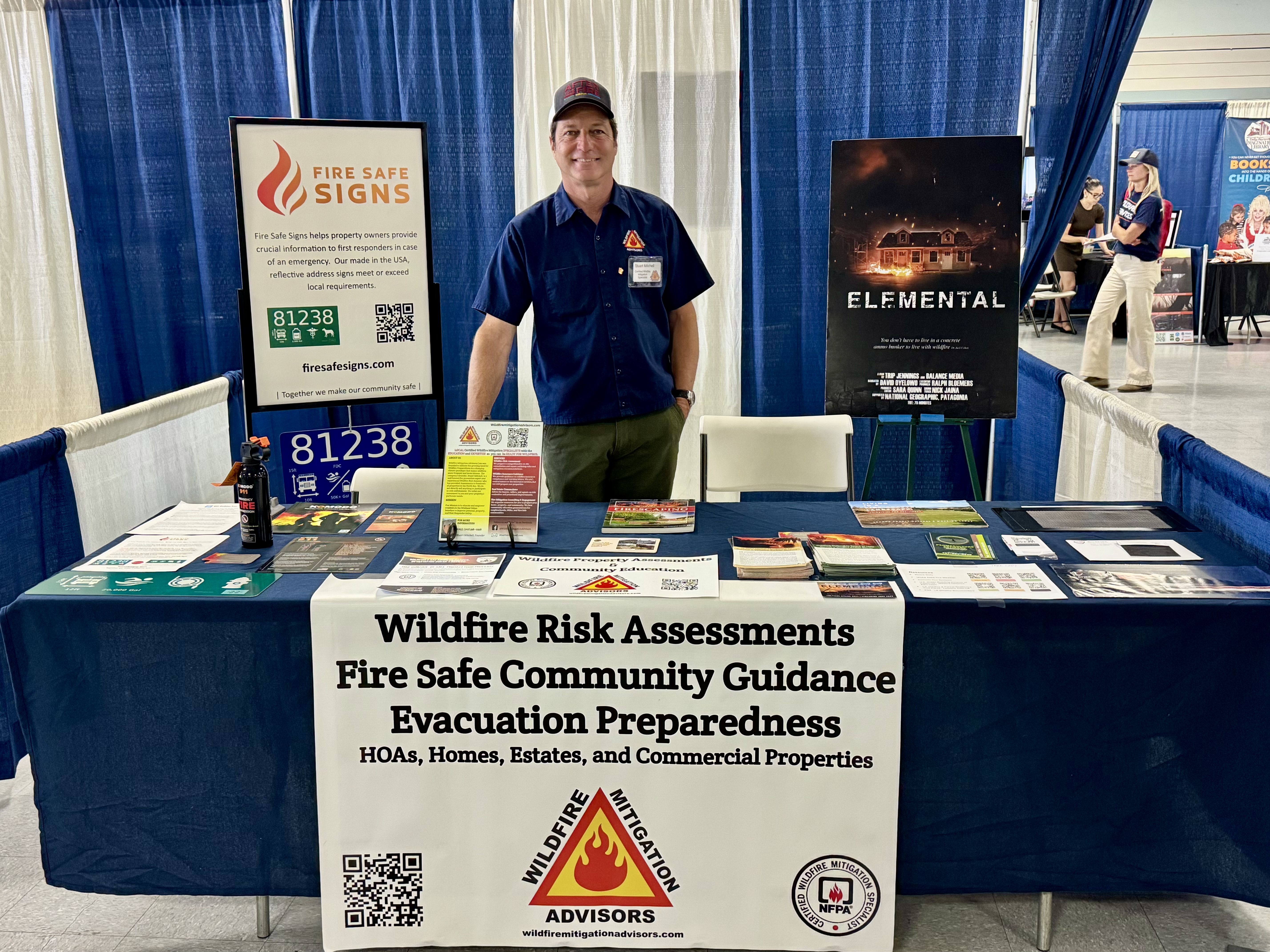 Stuart Mitchell at the Wildfire Mitigation Advisors booth at a community event, with WMA branding, Elemental documentary poster, and Fire Safe Signs display